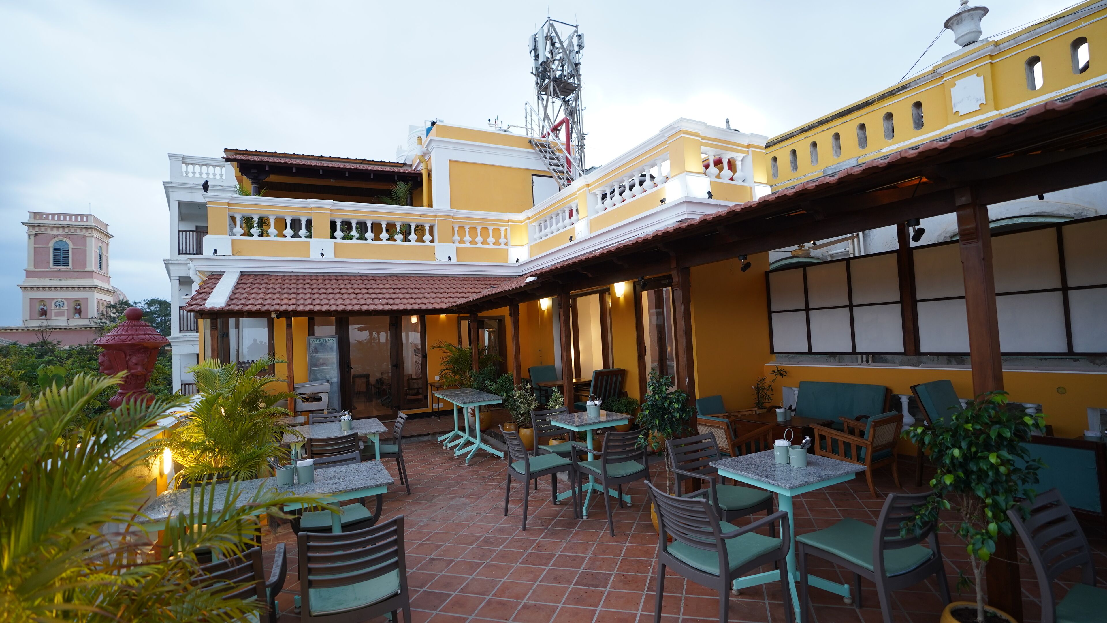 An outdoor patio area with multiple tables and chairs, a building with yellow and white walls, and a clock tower in the background.