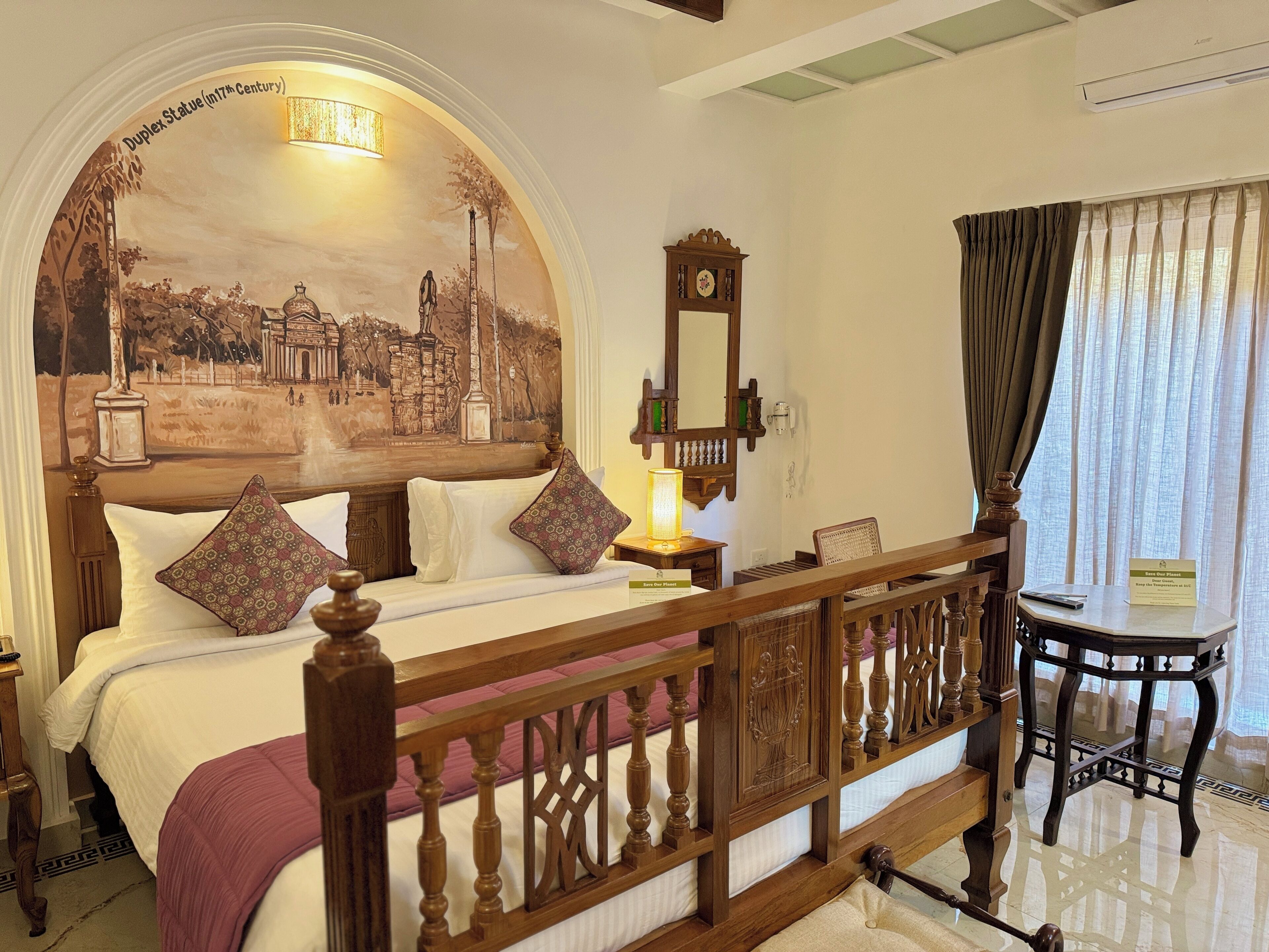 A hotel room with a bed, a wooden headboard, a nightstand, a lamp, a mirror, a window with curtains, and a small table.