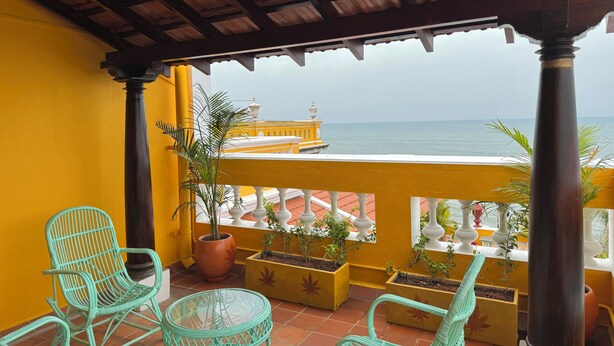 A balcony with wicker chairs, potted plants, and a view of the sea and a yellow building. - La Plage SeaView Suites (Puducherry)