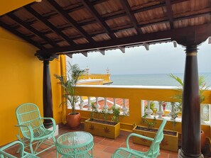 Ein Balkon mit Korbstühlen, Topfpflanzen und Blick auf das Meer sowie ein gelbes Gebäude.