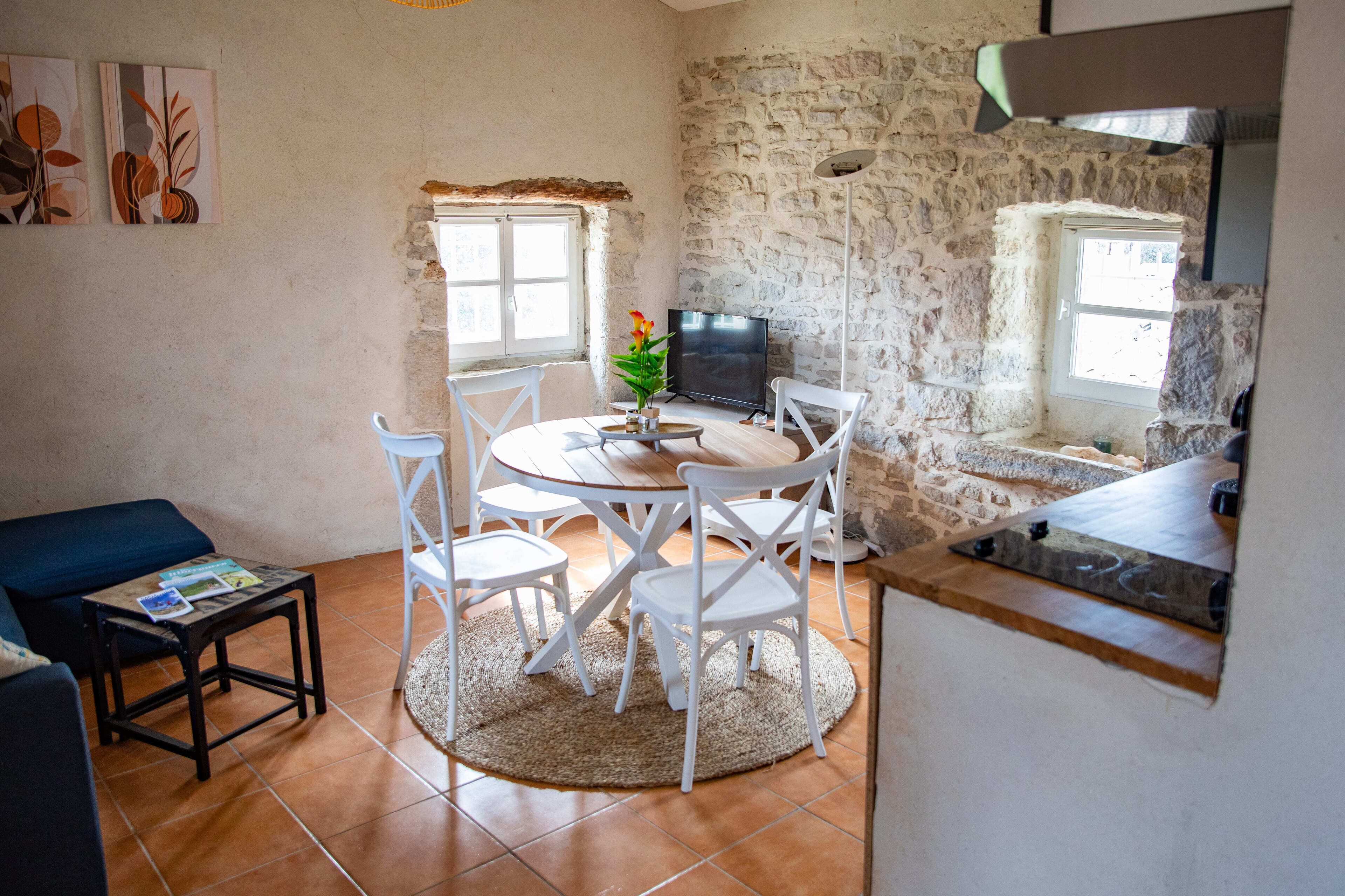 A cozy living room with a round table, white chairs, a TV, and a stone wall.
