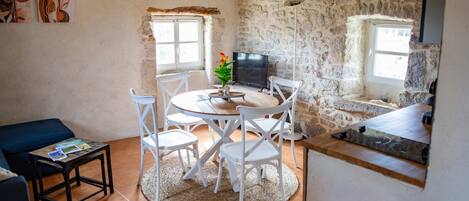 A cozy living room with a round table, white chairs, a TV, and a stone wall.