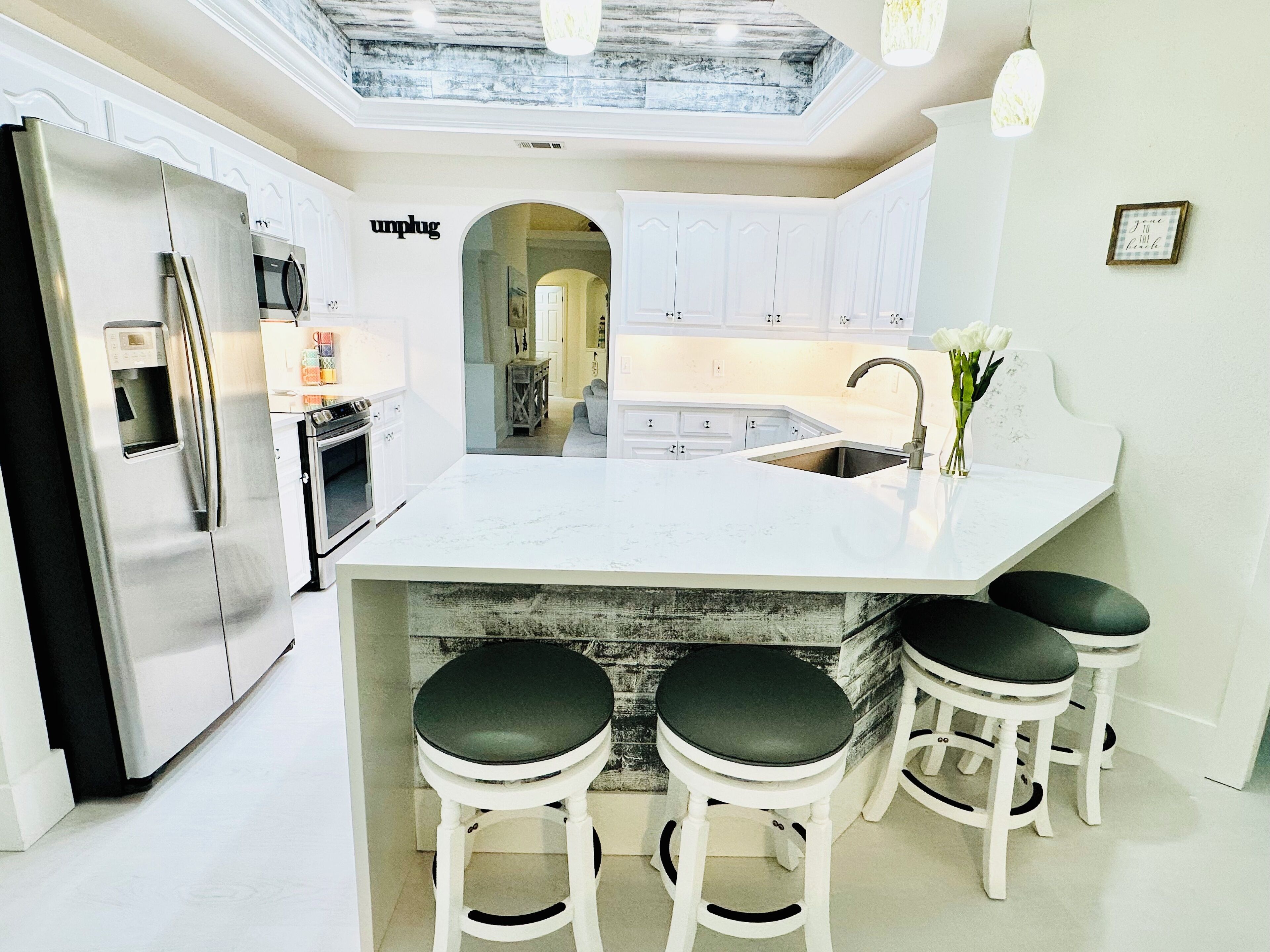 A modern kitchen with white cabinetry, a stainless steel refrigerator, a built-in oven, a marble countertop, a sink, and three bar stools with black seats.