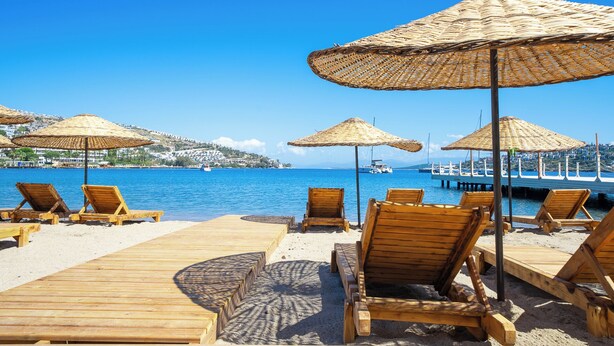 A beach with wooden lounge chairs and thatched umbrellas, a wooden boardwalk, and a view of boats and buildings across the water. - Mare Alba (Bodrum)