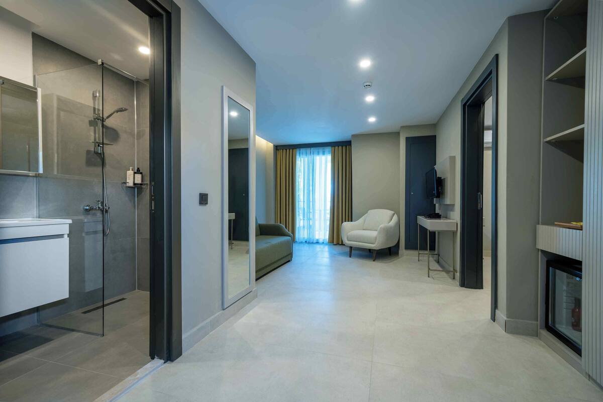 A modern hotel room with a glass shower, a sofa, a chair, a small table, and a fireplace.
