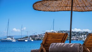 A beach with wooden lounge chairs and a woven umbrella. - Mare Alba (Bodrum)