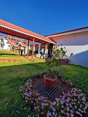 A building with a red roof and white walls, surrounded by a garden with pink flowers and a central planter. - Welbeck Vintage by Oxygen Resorts (Ooty)