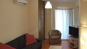 Una sala de estar con un sofá oscuro, una mesa de centro clara, un sillón color beige, un televisor sobre un mueble bajo y una lámpara.