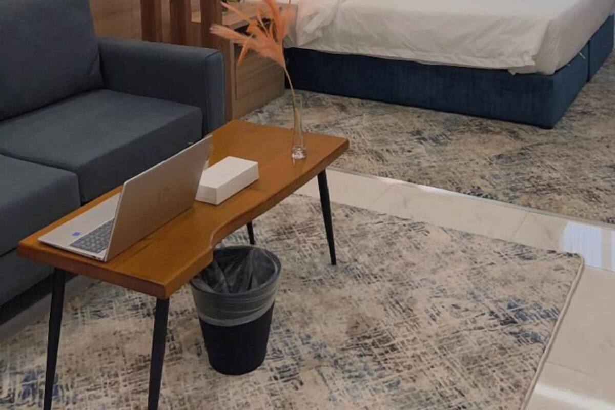 A hotel room with a bed, a sofa, a table with a laptop, and a trash bin.