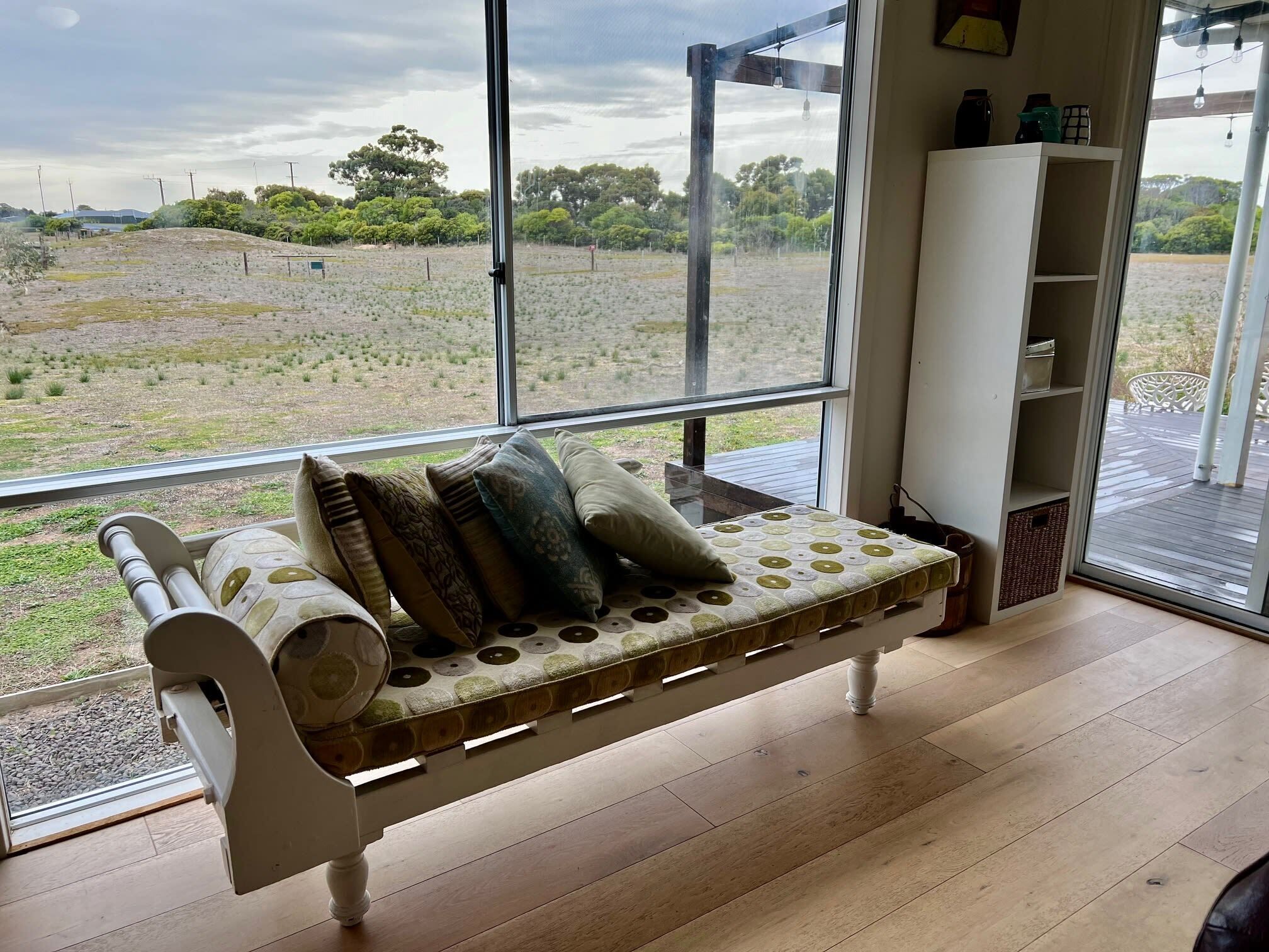 A room with a large window, a wooden bench with cushions, a white cabinet, and a view of a grassy field.