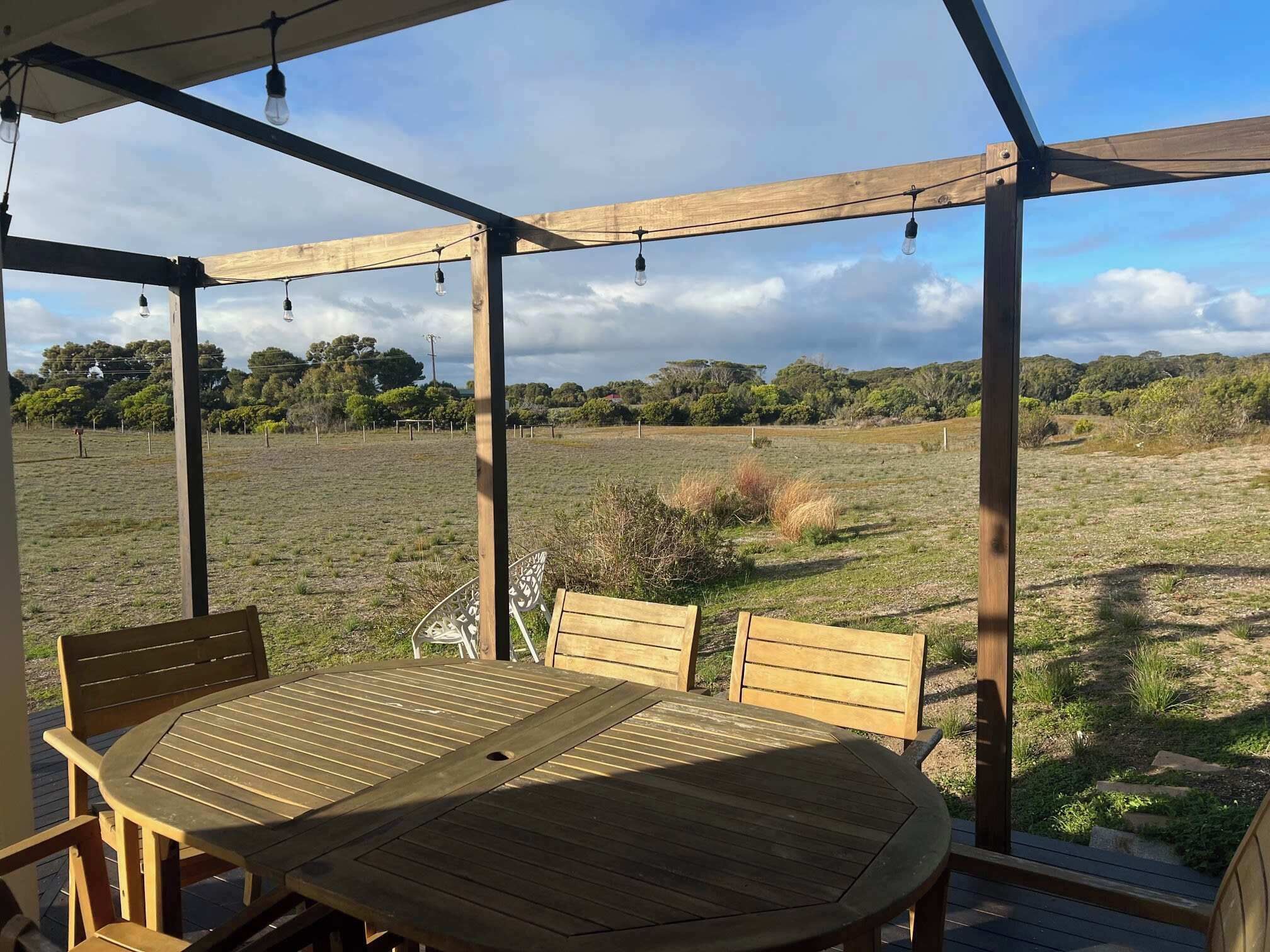 An outdoor wooden deck with a table and chairs, a pergola with string lights, and a view of a grassy field and trees.