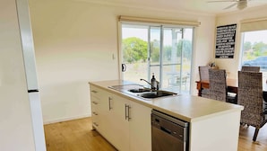 A modern kitchen with a dishwasher, a window, and a dining area with wicker chairs. - Ramblers Croft- WiFi, Pet friendly outside (Robe)