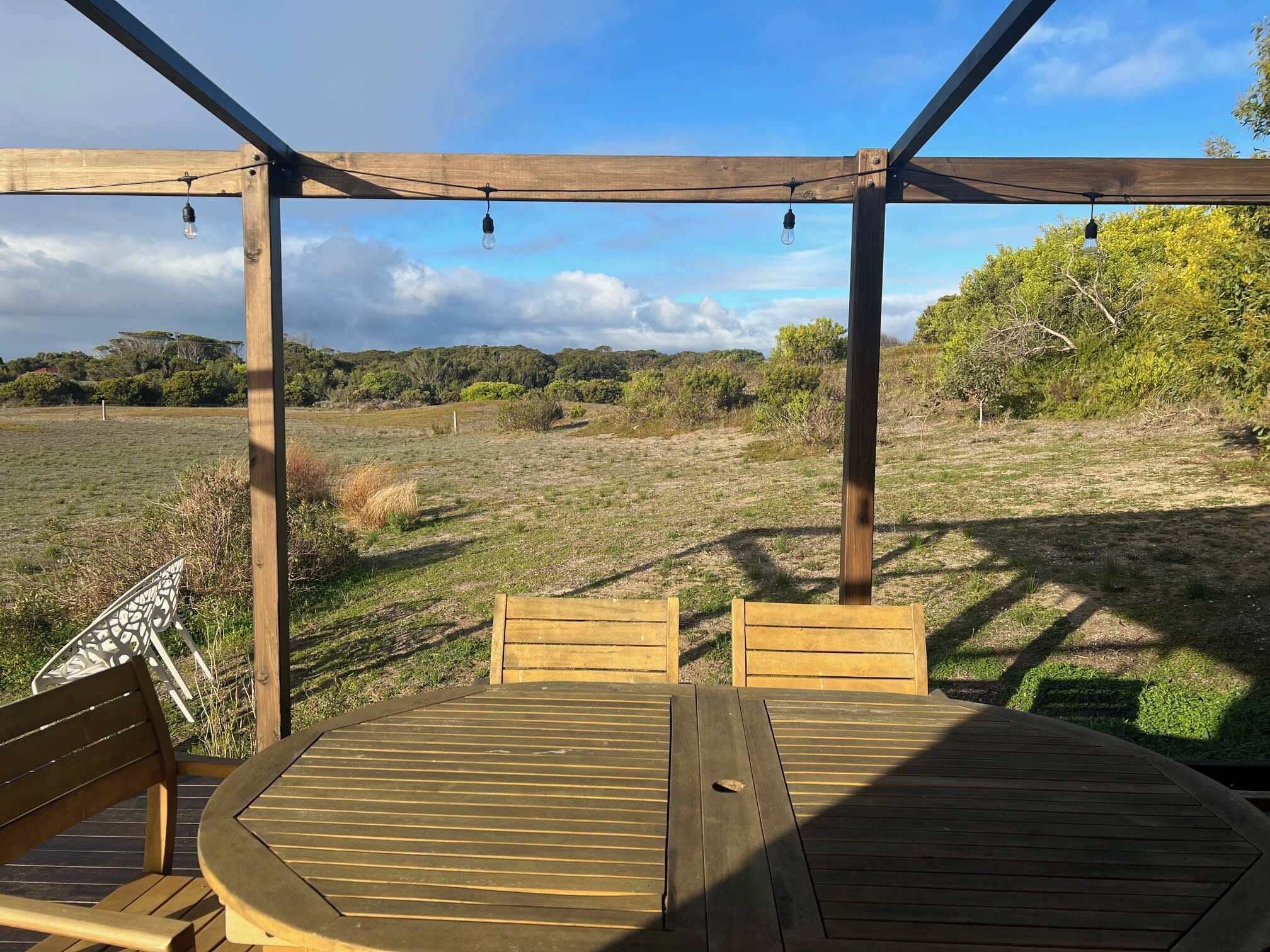 Une terrasse en bois avec une table et des chaises, entourée d’une pergola en bois ornée de guirlandes lumineuses, donnant sur un champ verdoyant parsemé d’arbres.
