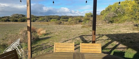 Une terrasse en bois avec une table et des chaises, entourée d’une pergola en bois ornée de guirlandes lumineuses, donnant sur un champ verdoyant parsemé d’arbres.