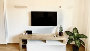 A modern living room with a flat-screen TV mounted on the wall, a wooden entertainment unit with a TV stand, and two potted plants. - Fully Renovated Accomodation (Fiumicino)