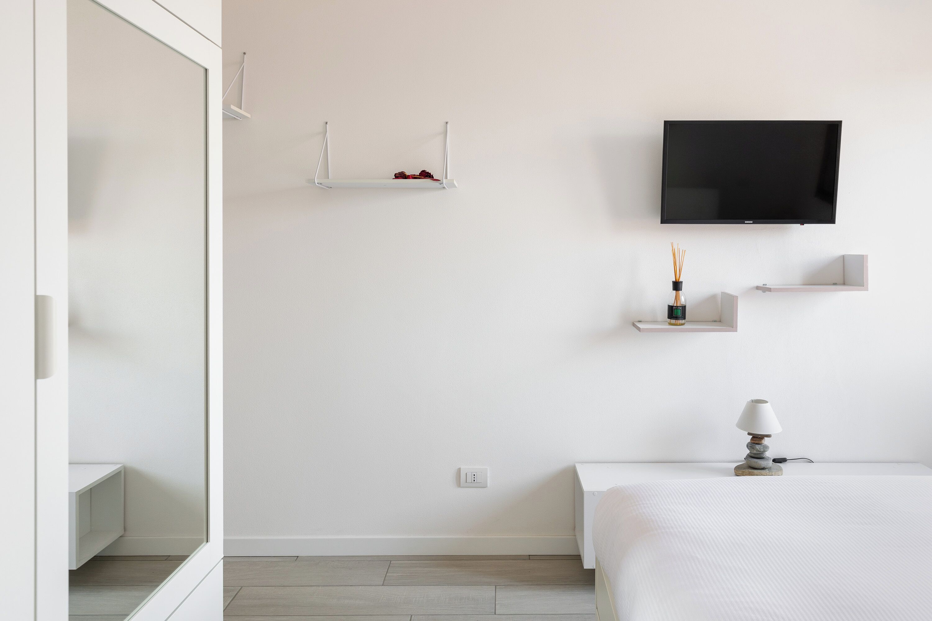 Une chambre à coucher au style minimaliste, avec un lit, un téléviseur fixé au mur et une table de chevet avec une lampe.