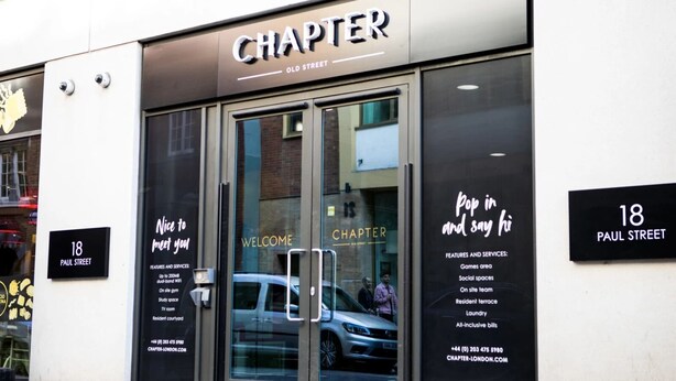 Voici l’extérieur d’un bâtiment appelé « CHAPTER », situé dans la rue Paul. Il se distingue par une grande entrée en verre sur laquelle est affiché un panneau « BIENVENUE », ainsi qu’un panneau indiquant les différents équipements disponibles, tels qu’un espace de jeux, des espaces de convivialité et une équipe présente sur place.