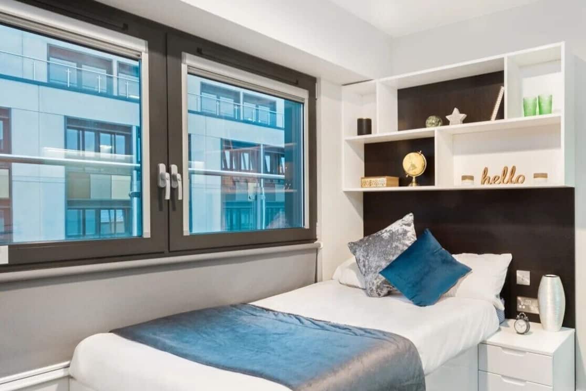 A modern bedroom with a bed, bedside tables, a nightstand, a shelf with decorative items, and a window with a view of buildings.