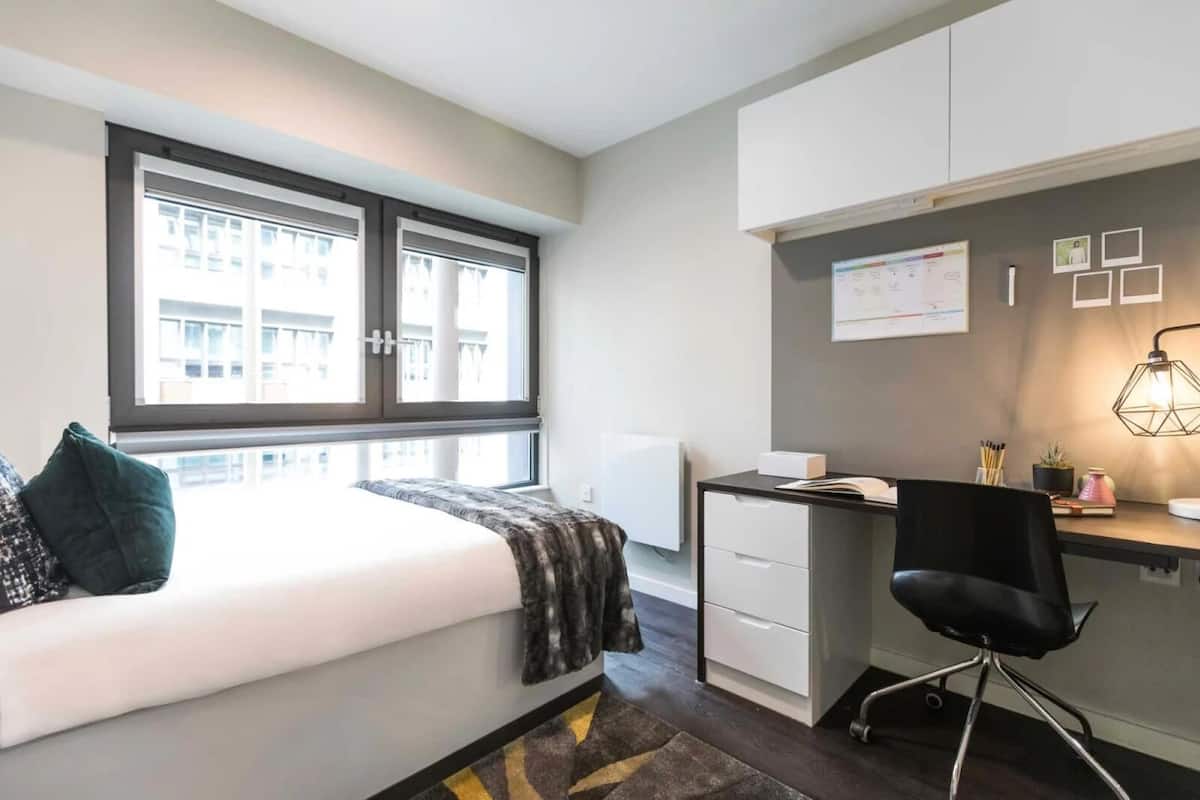 A modern bedroom with a bed, a desk, and a chair. There is a window with blinds, a radiator, and a wall-mounted shelf with books and a lamp.
