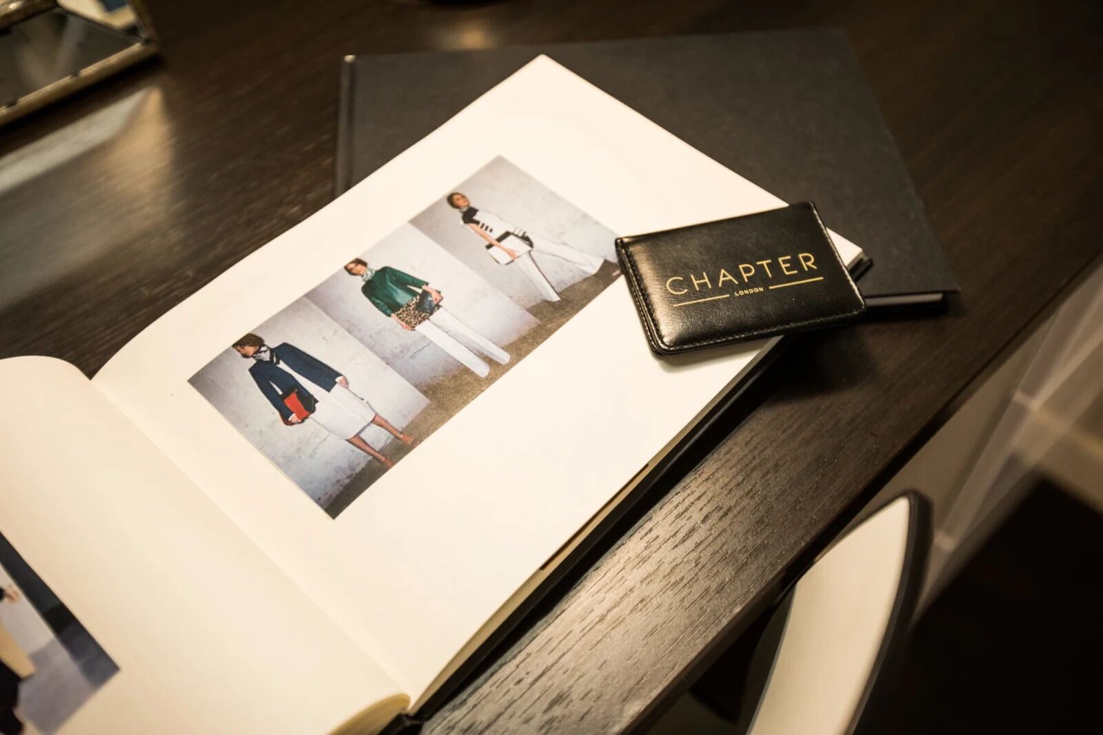 Un libro abierto con una página que muestra modelos de moda, un libro de tapa de cuero negro titulado "CHAPTER LONDON" sobre una superficie de madera.