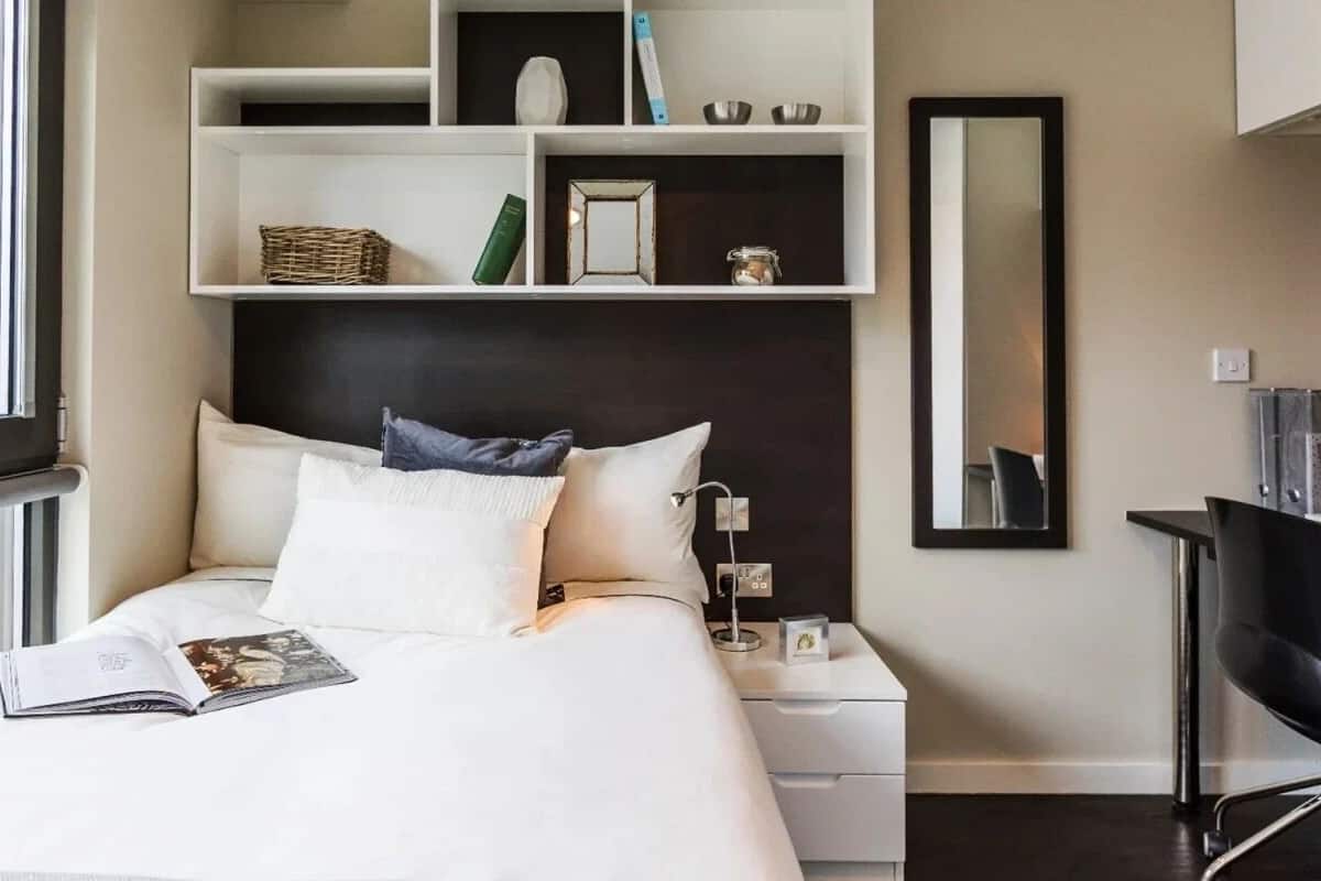 A modern hotel room with a neatly made bed, a bedside table with a lamp, a small shelf with decorative items, a desk with a chair, and a large window providing a view of the city.