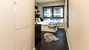 Un couloir étroit mène à une chambre dotée d’un lit, d’étagères intégrées et offrant une vue sur les bâtiments grâce à de grandes fenêtres.