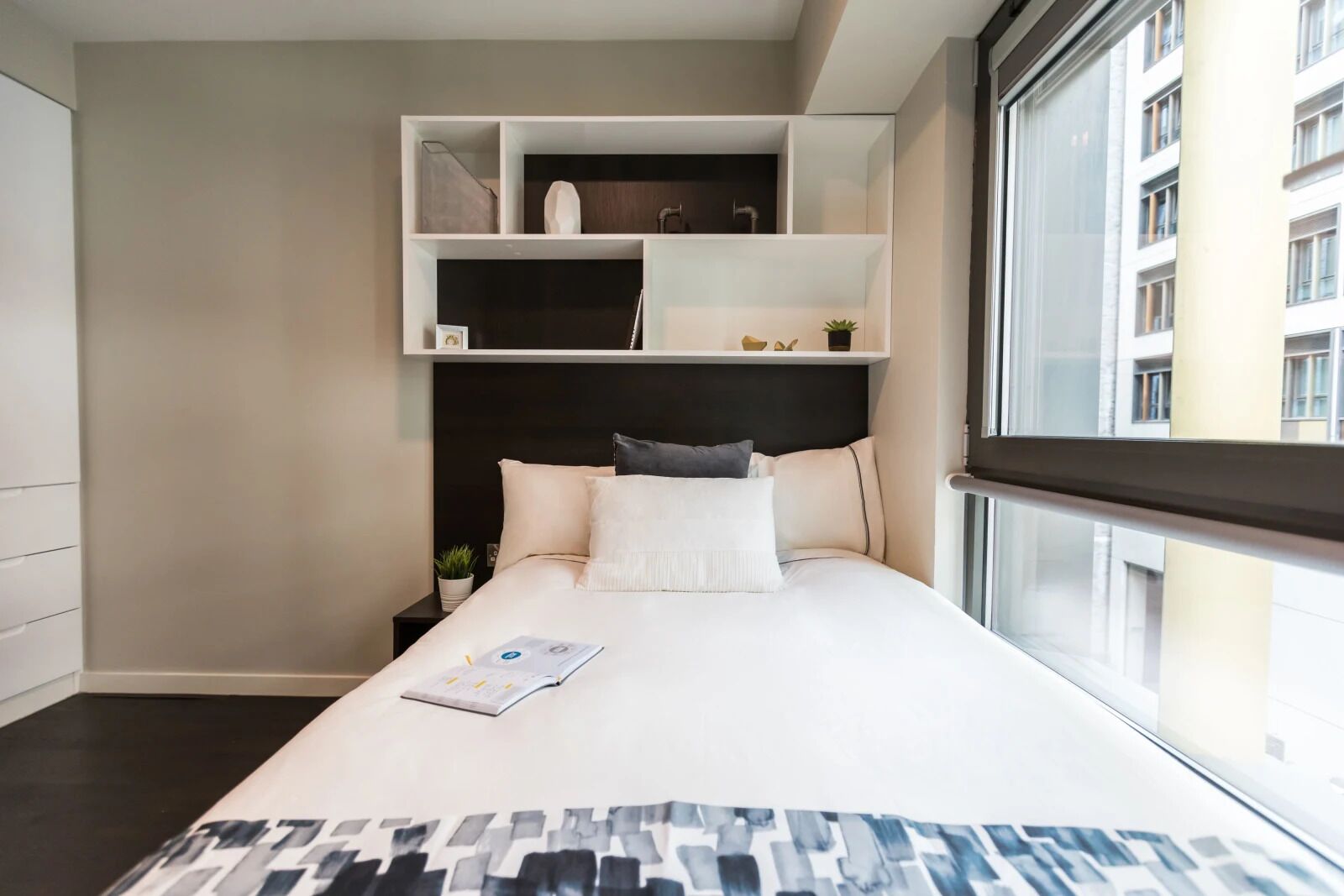 Habitación de hotel moderna con una cama grande, estanterías empotradas y vistas a un paisaje urbano a través de un ventanal amplio.