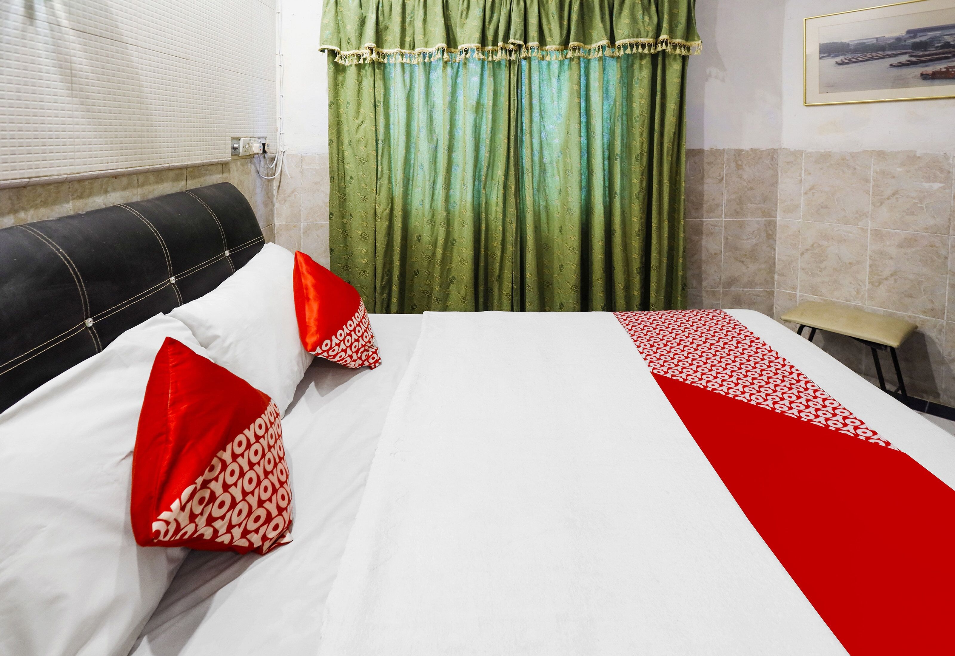 Una habitación de hotel con cama, almohadas, cabecero, ventana con persianas y una cortina verde, además de un cuadro colgado en la pared.