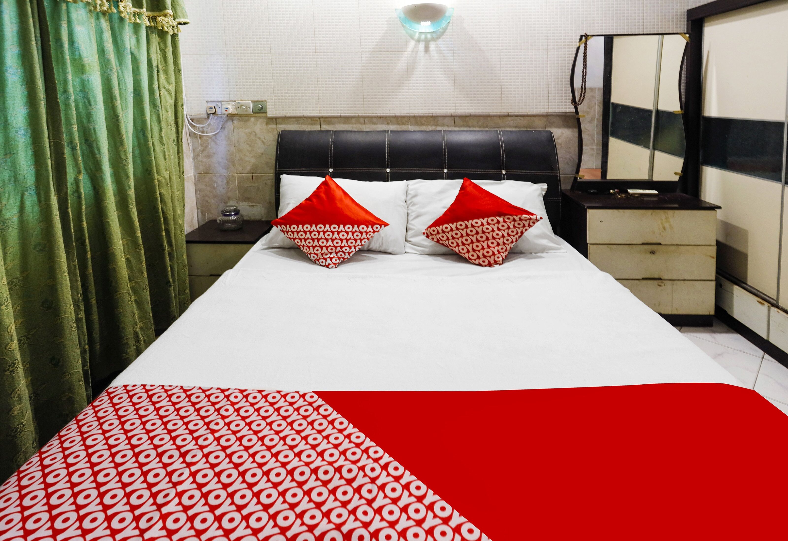 A hotel room with a bed, two pillows, a nightstand, a mirror, and a green curtain.