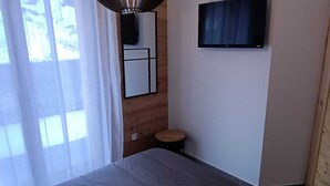 A modern bedroom with a bed, a mounted TV, a small stool, and a window with sheer curtains. - Apartment Vars, 2 bedrooms, 7 persons (Vars)