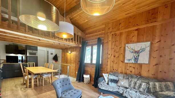 A cozy cabin interior with a wooden dining area, a gray tufted chair, a blue sofa with patterned cushions, and a small round table.