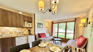 Eine gemütliche Küche mit Holzausstattung, einer Mikrowelle und einer Kaffeemaschine. Ein Essbereich mit Tisch für Zwei, komplett mit Tellern, Gläsern und Besteck. Das Zimmer hat ein Fenster mit Vorhängen und Blick ins Grüne.