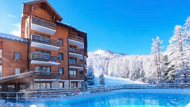 Ein Hotel mit Pool, Holzfassade, Balkonen und einem verschneiten Berg im Hintergrund.
