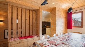 A cozy wooden cabin with a dining area, a bench with a red cushion, and a kitchen visible through an open door.