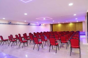 A conference room with rows of red chairs, a stage with a golden curtain, and ceiling lights. - Shri Valli Resdiency (Chennai)