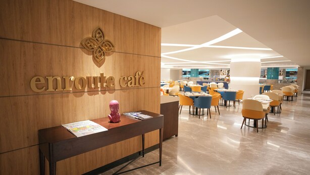 A reception area with a wooden wall and a sign reading "enroute cafe". There are tables and chairs arranged in the background. - Transit by Encalm - Nap and Shower (Hyderabad)