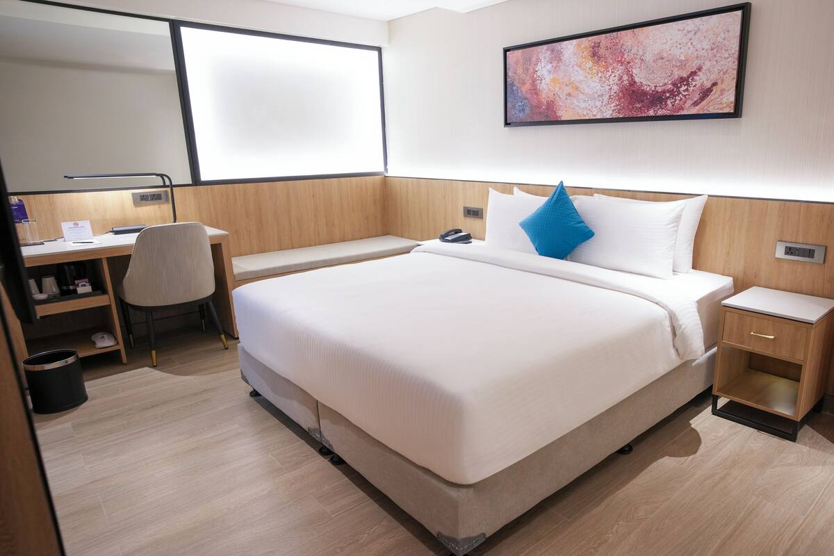 A hotel room with a large bed, a desk with a chair, a small bedside table, a trash bin, a wall-mounted phone, and a painting.