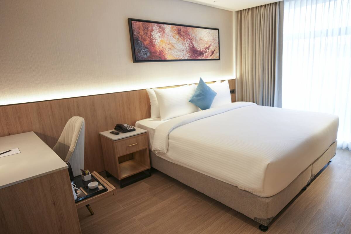 A hotel room with a large bed, a desk, a chair, a nightstand with a phone, and a painting on the wall.