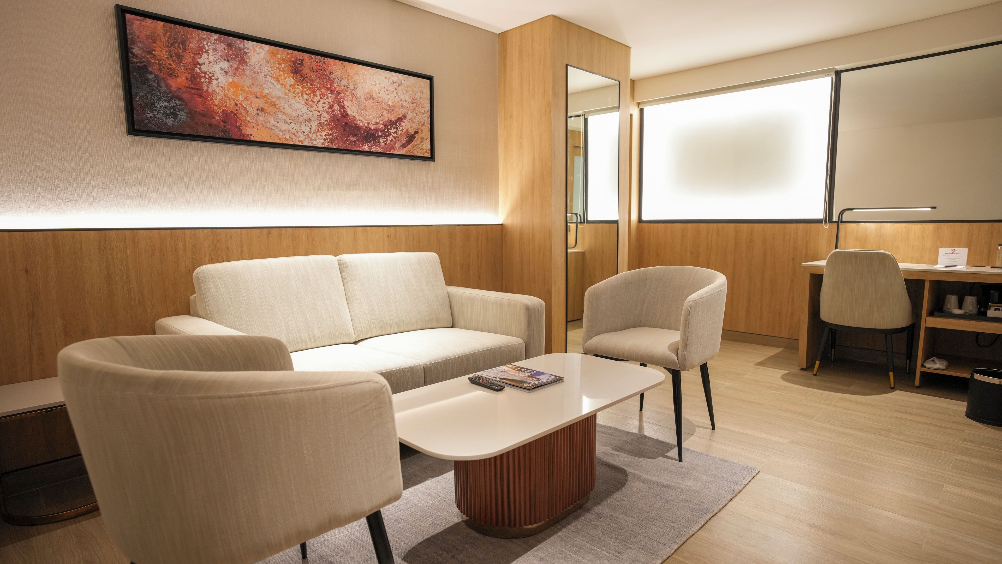 A modern hotel room with a sofa, armchairs, a coffee table, a desk, and a large abstract painting on the wall.