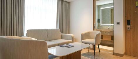 Una habitación de hotel moderna con sofá, dos sillones, una mesita de centro y un baño visible a través de un ventanal.