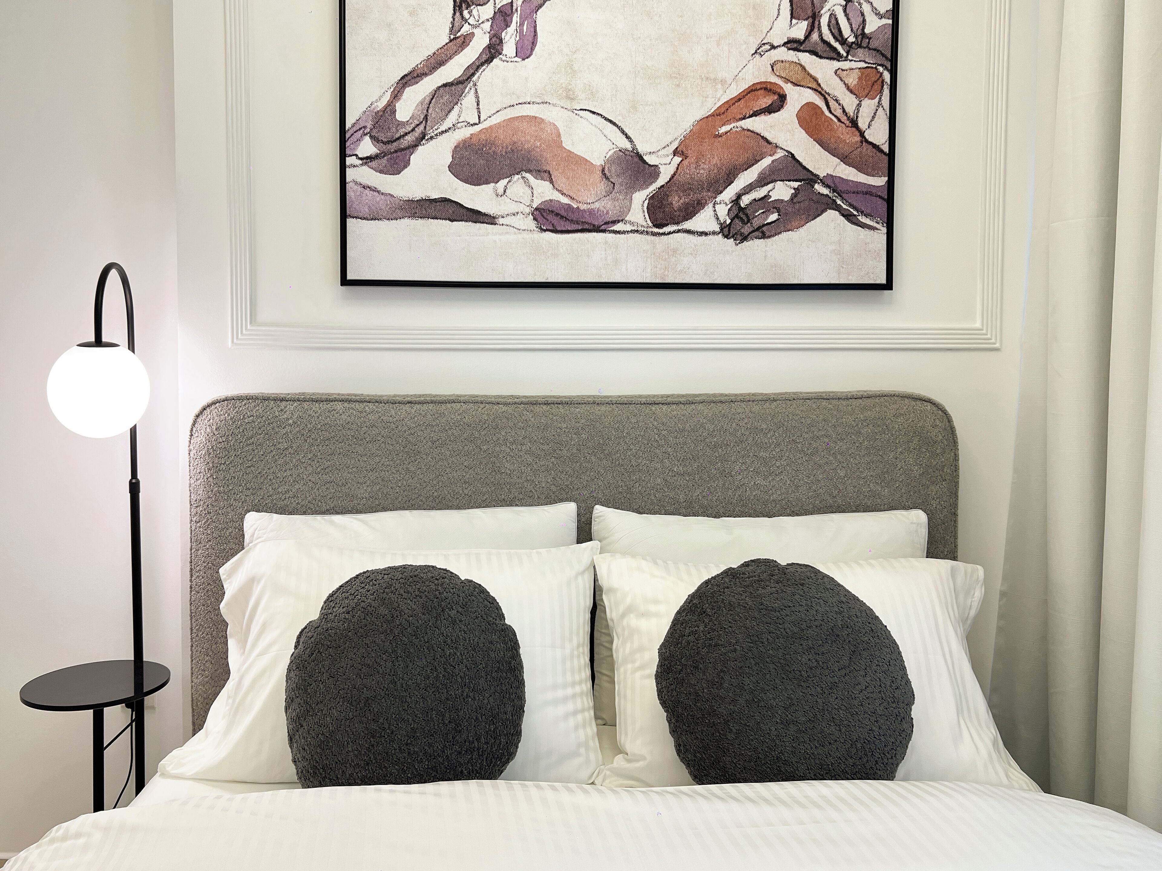 Ein ordentlich bezogenes Bett mit zwei weißen Kissen und zwei großen schwarzen Zierkissen, eine moderne Stehlampe und ein gerahmtes abstraktes Gemälde über dem Bett.