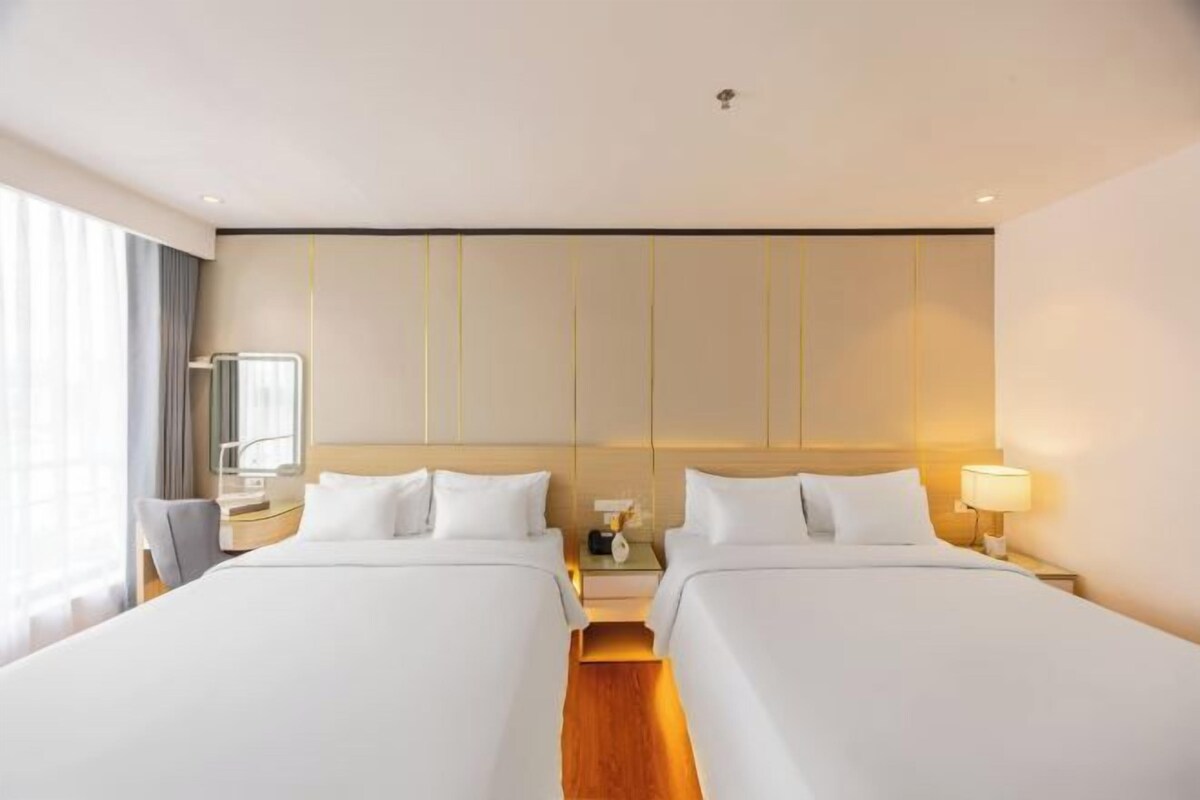 A hotel room with two beds, a bedside table with a lamp, a nightstand, a mirror, and a window with curtains.