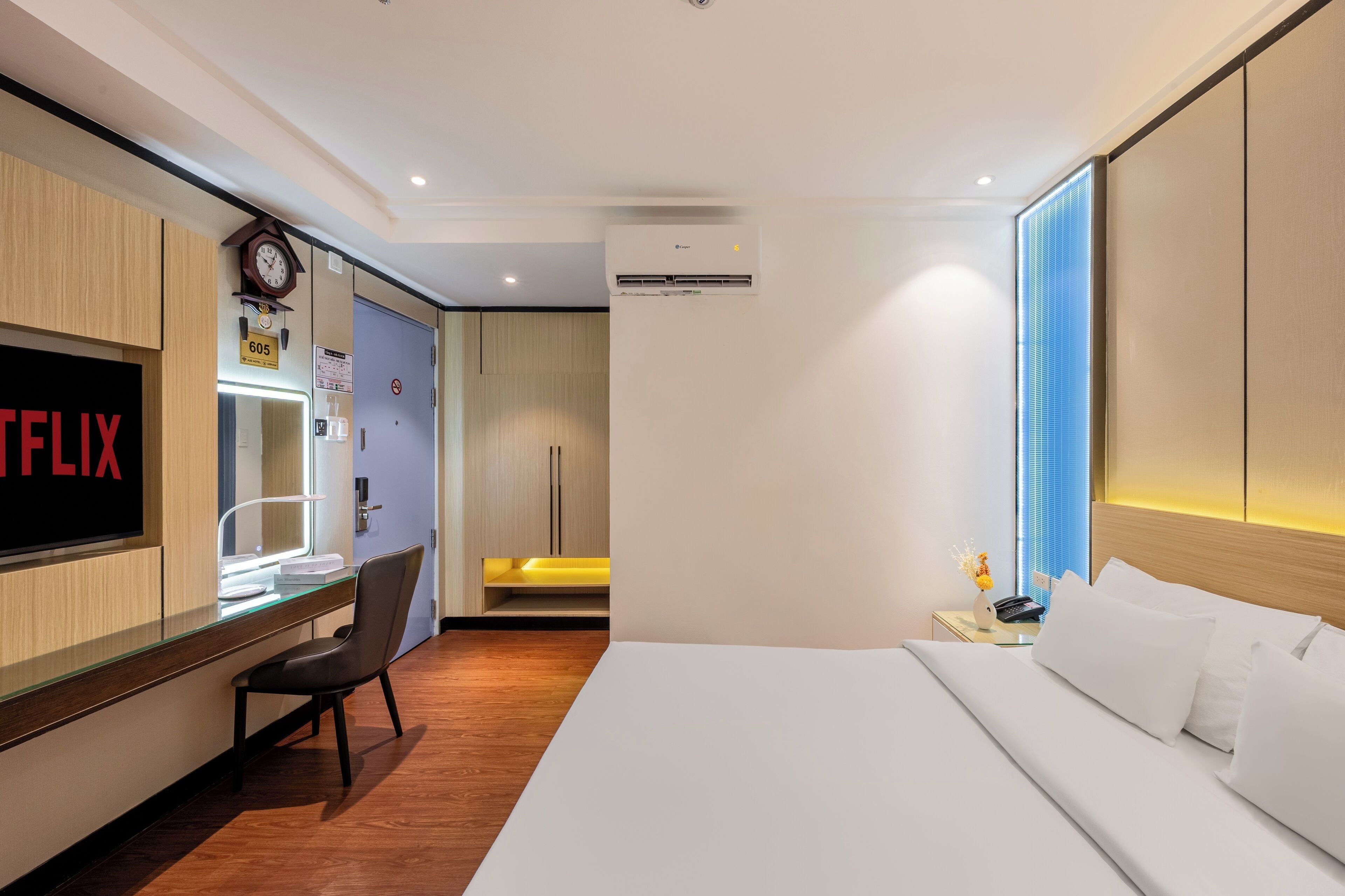 A hotel room with a bed, a desk with a chair, a TV, an air conditioner, and a window with blinds.