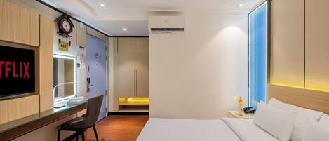 A hotel room with a bed, a desk with a chair, a TV, an air conditioner, and a window with blinds.