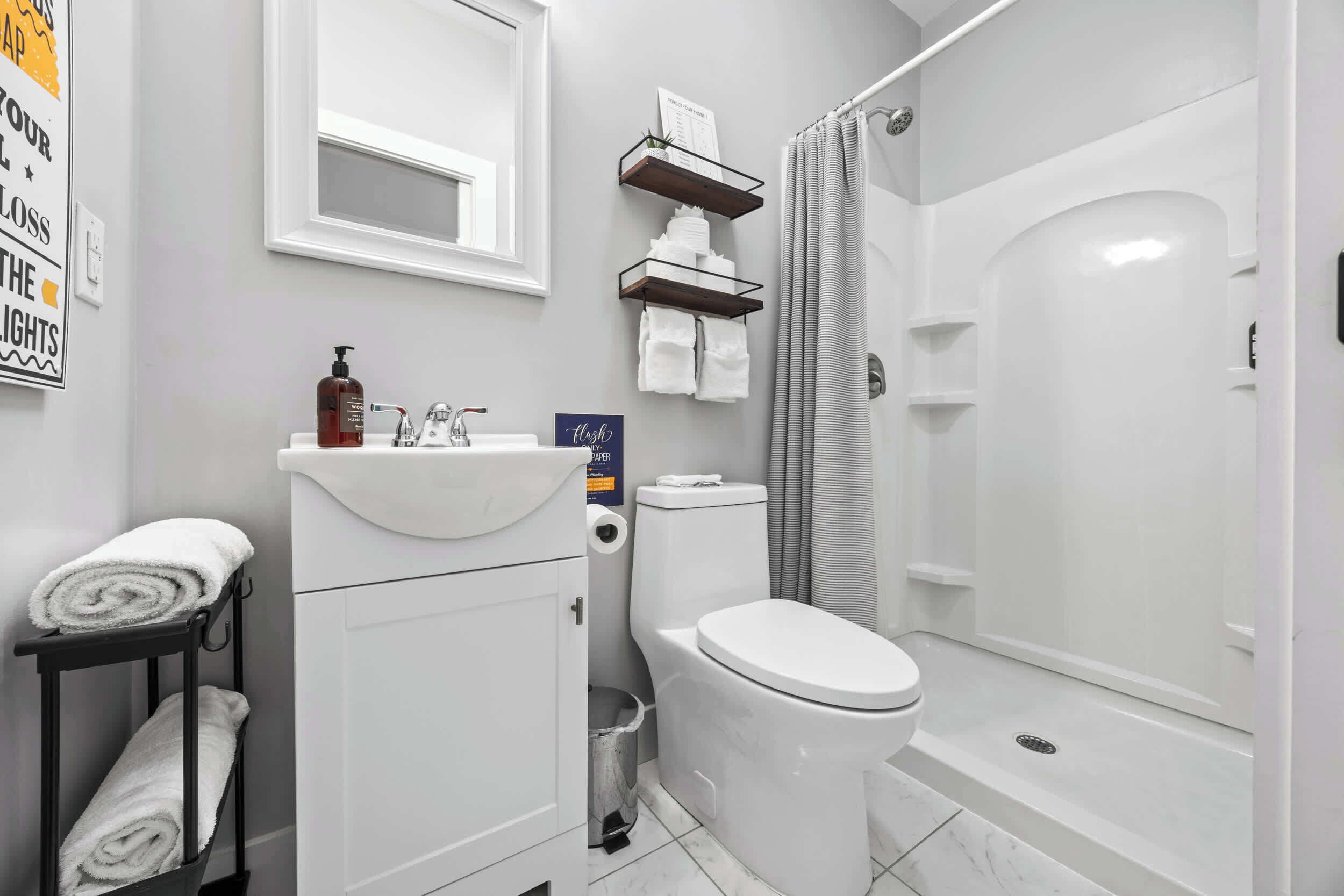 A modern bathroom with a white sink, toilet, and shower. There are shelves above the toilet and a towel rack beside the sink.