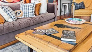 Een gezellige woonkamer met een bruine bank, gestreepte kussens, een houten bijzettafeltje met een boek en onderzetters, en een keuken in de achtergrond.