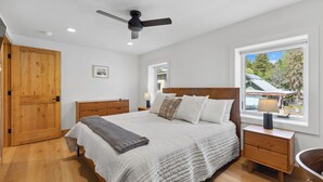 Un dormitorio con una cama grande, cabecera de madera, ventilador de techo, ventana con vista a árboles y un armario de madera.