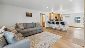 Una sala moderna con un sofá gris, una zona de cocina con taburetes de bar y un suelo de madera.