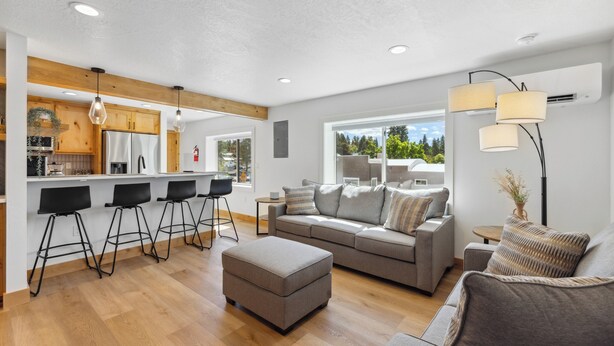 A modern living room with a grey sofa, ottoman, and black bar stools. A kitchen with wooden cabinets and a large window is visible in the background. - Downtown Loft 205 - Downtown McCall condo next to Payette Lake (McCall)