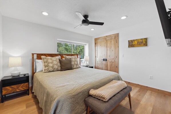 Une chambre à coucher comprenant un lit, une table de chevet, une lampe, un banc, un ventilateur de plafond, une fenêtre et une armoire en bois.